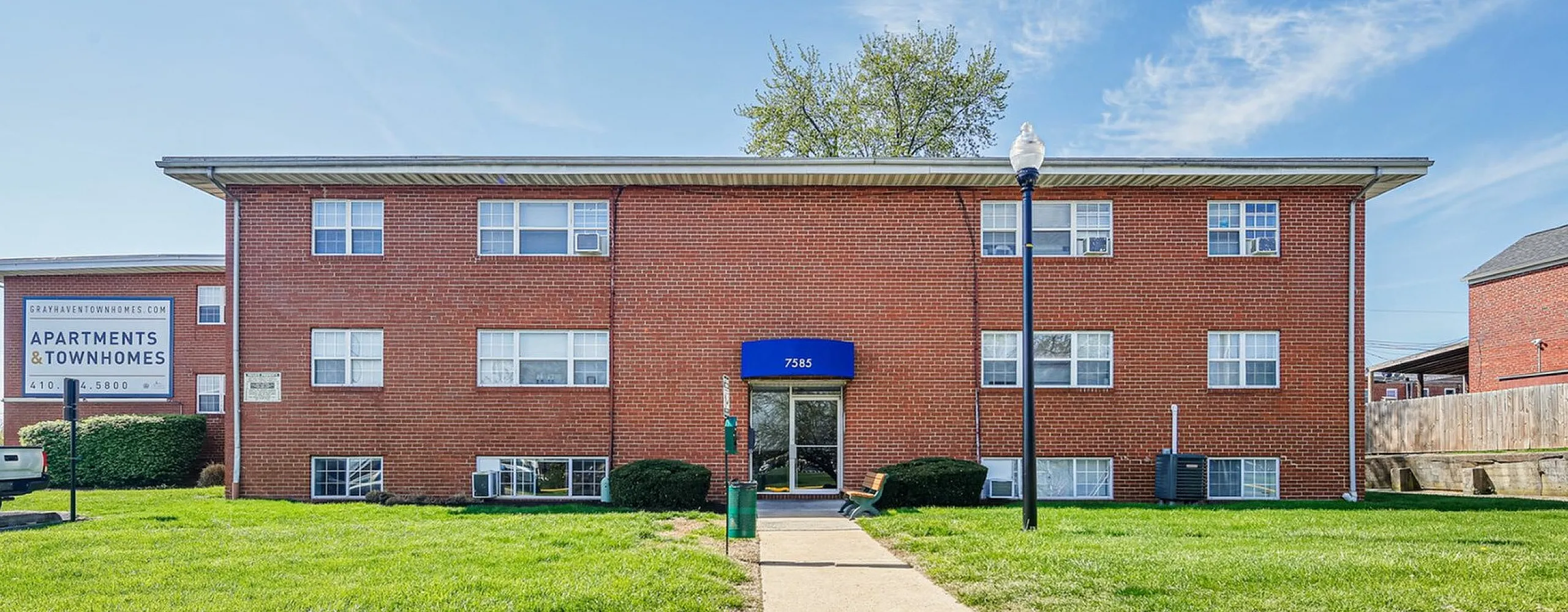 Charming red brick apartment complex with well-maintained greenery and a welcoming entrance under a bright blue sky.
