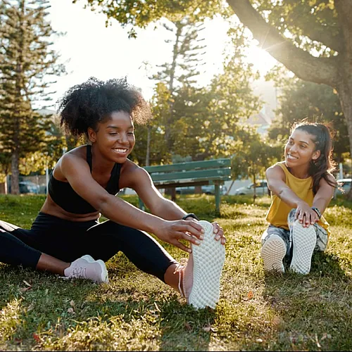 Two young women enjoying a sunny day, stretching on a lush green lawn in a peaceful park setting.