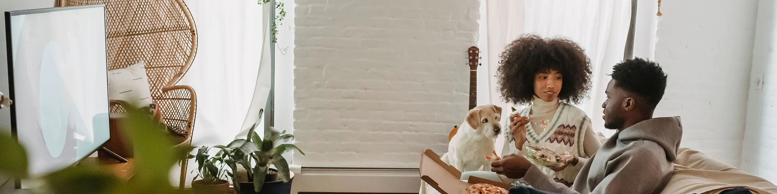 Cozy apartment living room with modern decor, featuring a couple enjoying pizza and salad on a couch, a friendly dog, elegant wicker chair, and lush plants adding charm and warmth.