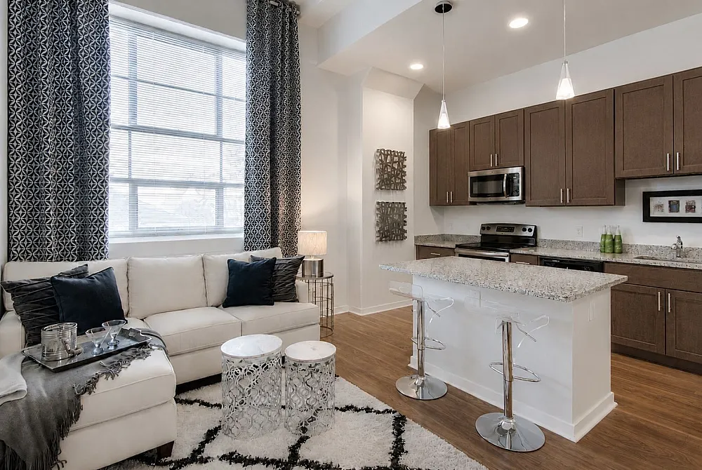 Bright and modern apartment living room with sleek kitchen, featuring elegant brown cabinets, stainless steel appliances, and a cozy white sofa under large patterned curtains.