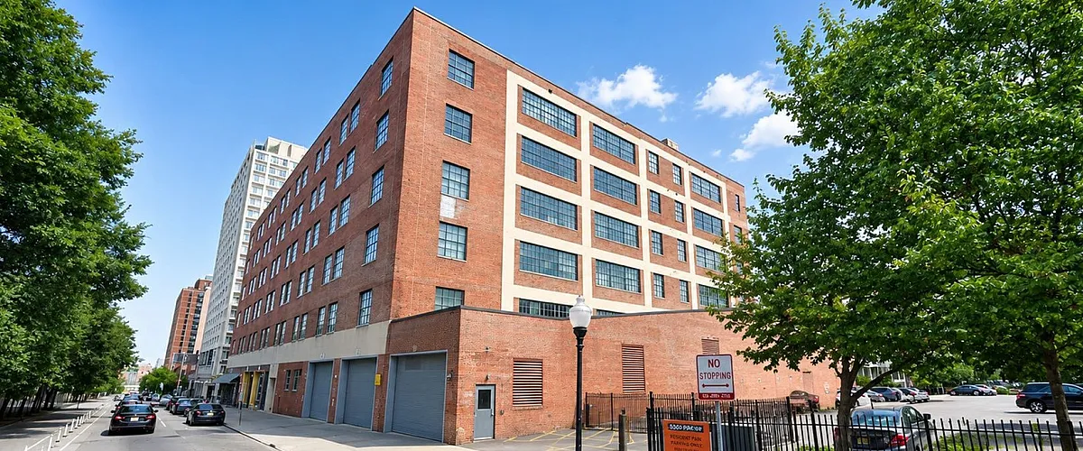 Charming urban streetscape featuring a classic brick apartment building with large windows, flanked by lush green trees and a vibrant blue sky.