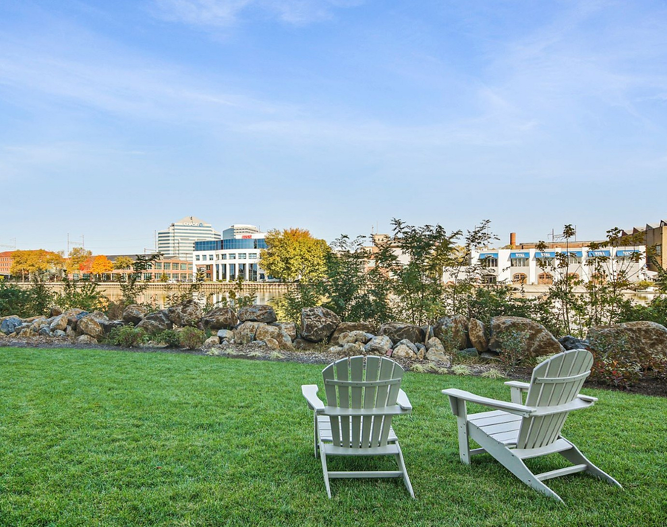 Scenic outdoor space with lush green grass, comfortable Adirondack chairs, and a backdrop of a river and cityscape.