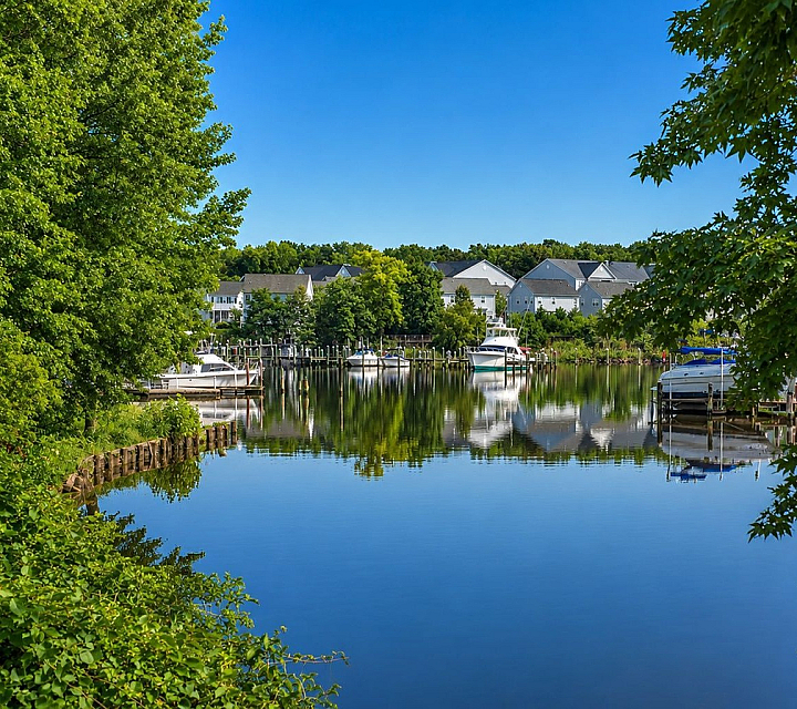 Serene waterfront view with charming townhouses, surrounded by lush greenery and tranquil waters reflecting the clear blue sky.