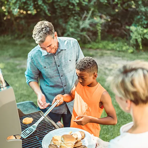 Joyful family barbecue in a lush backyard setting, featuring burgers and sausages on the grill under a sunny sky.