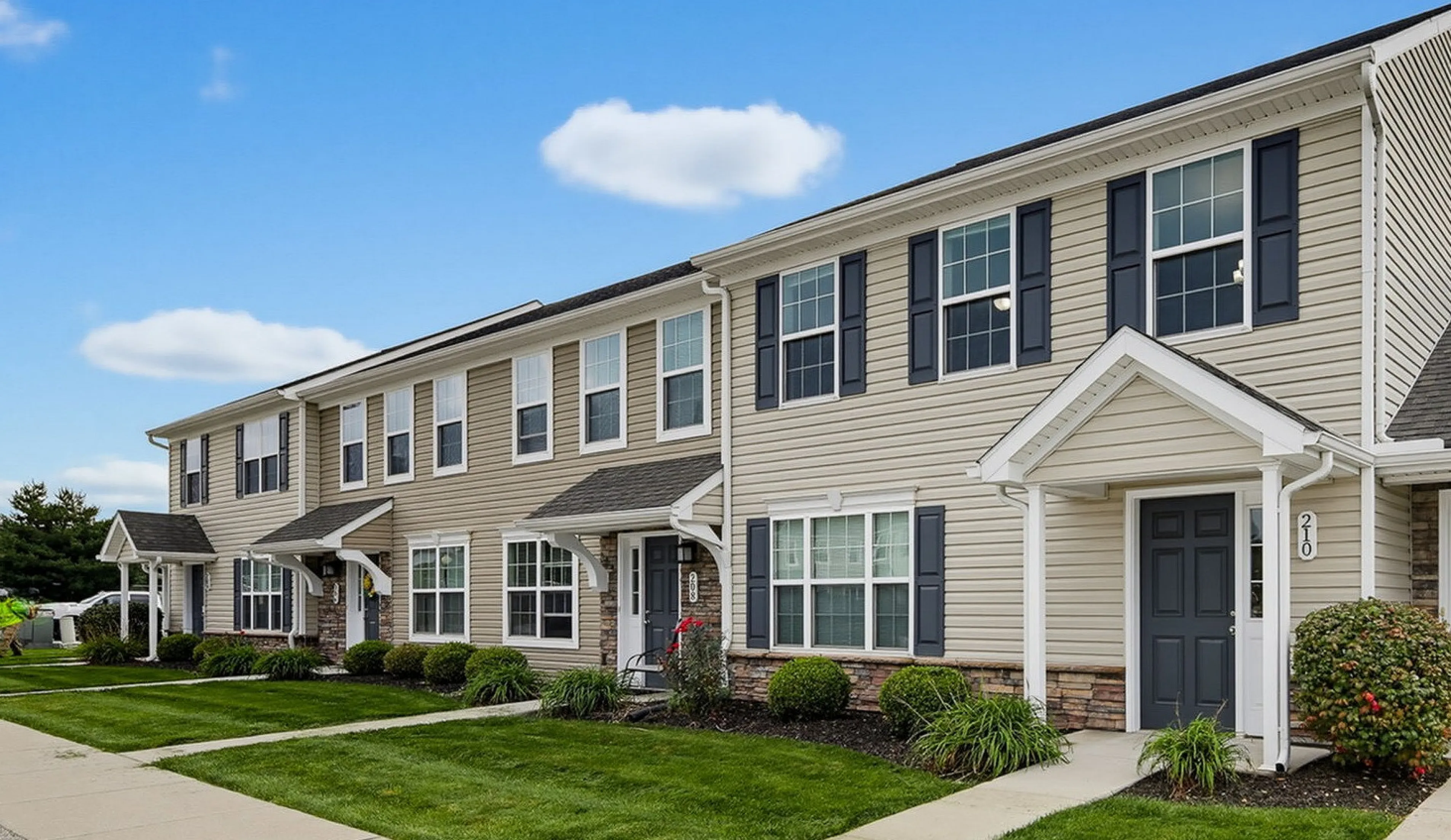 Charming two-story townhouses with landscaped lawns and welcoming porches under a bright blue sky.