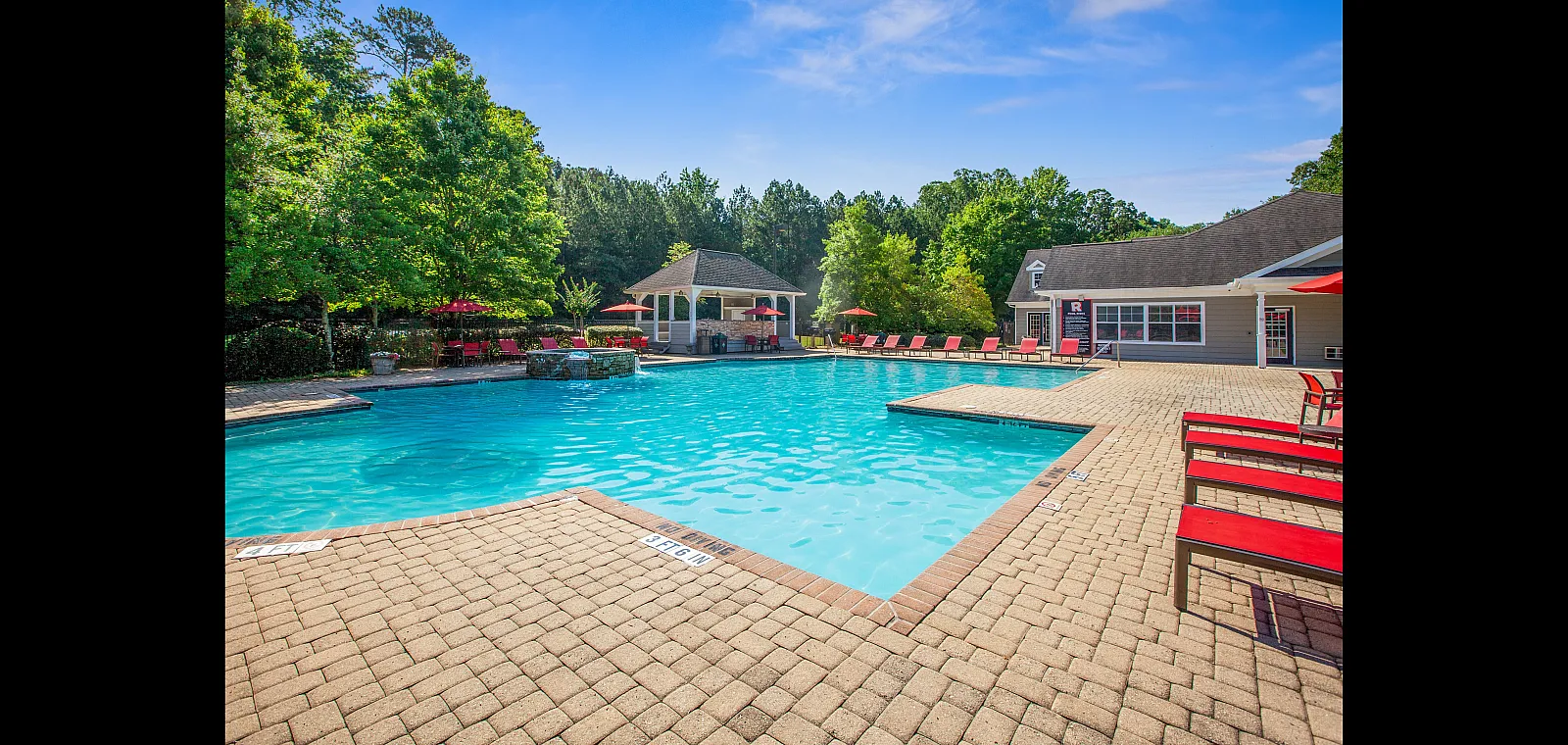 Inviting outdoor swimming pool area with red lounge chairs and lush greenery, perfect for relaxation under a clear blue sky.
