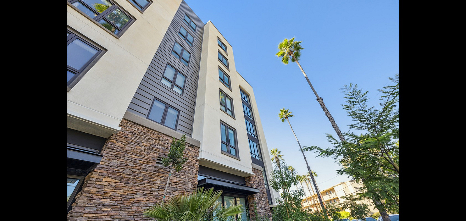 Modern apartment building with stylish stone facade surrounded by palm trees, set against a clear blue sky.