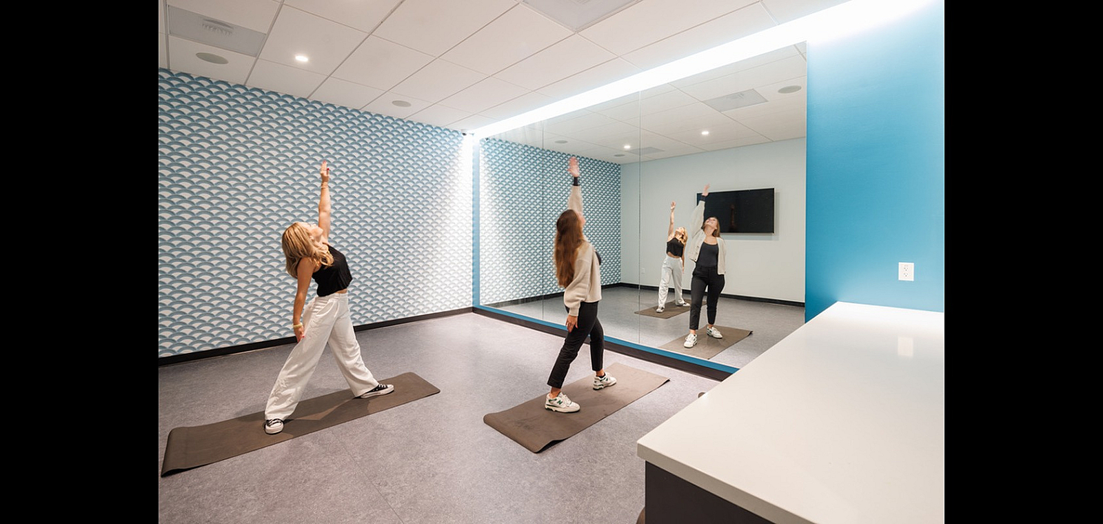 Modern fitness room with large mirrors and stylish wall pattern, featuring individuals practicing yoga in a bright and inviting space.