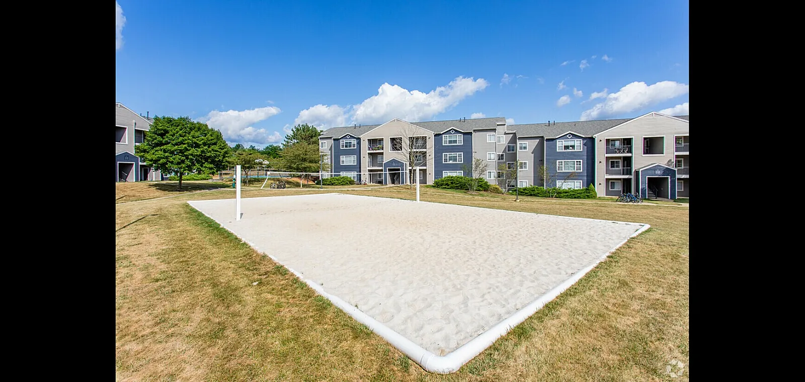 Charming apartment complex with modern facades, lush green surroundings, and an inviting sandy volleyball court under a clear blue sky.