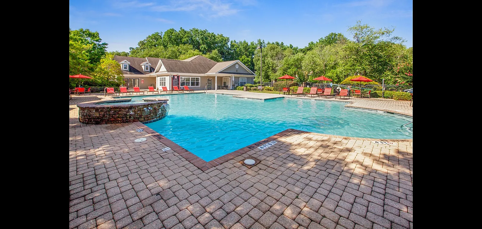 Inviting outdoor pool area with crystal-clear water, surrounded by cozy lounge chairs and vibrant red umbrellas, bordered by lush greenery.
