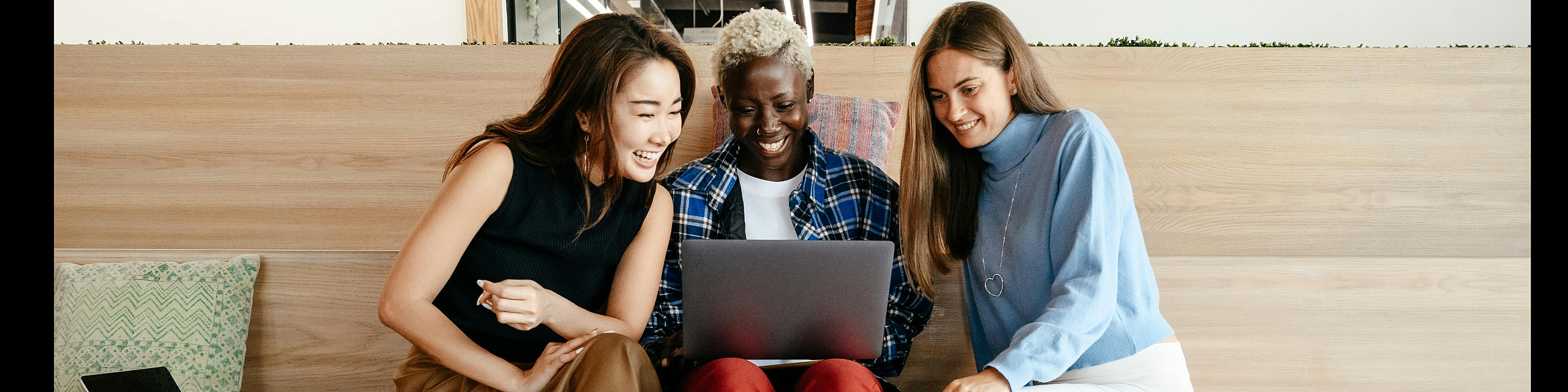 Joyful group of three friends sitting on modern wooden steps, engaging with a laptop, embodying a stylish and collaborative atmosphere.