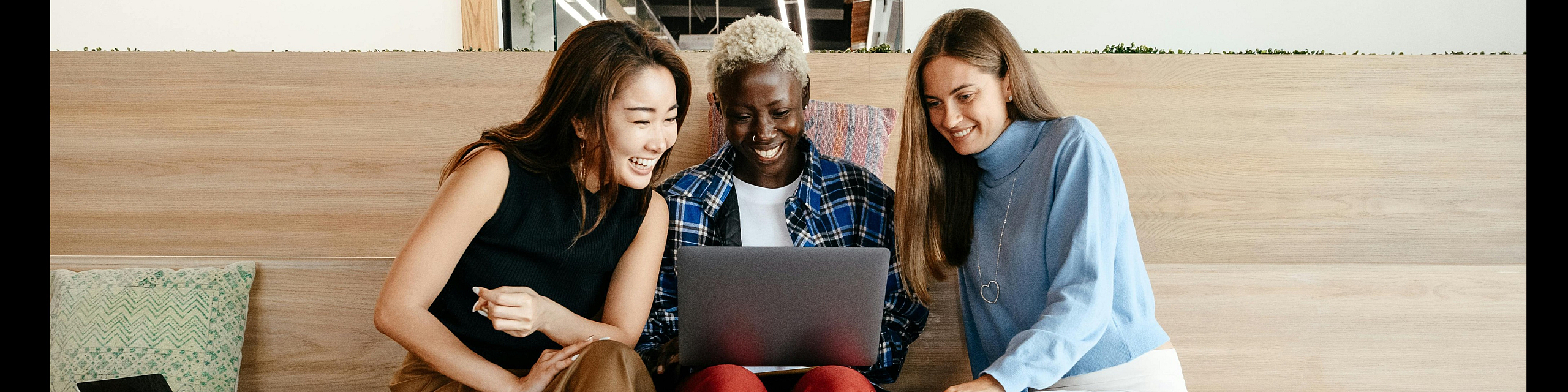 Three diverse individuals joyfully engaging with a laptop in a modern, inviting workspace with wooden steps and cozy cushions.