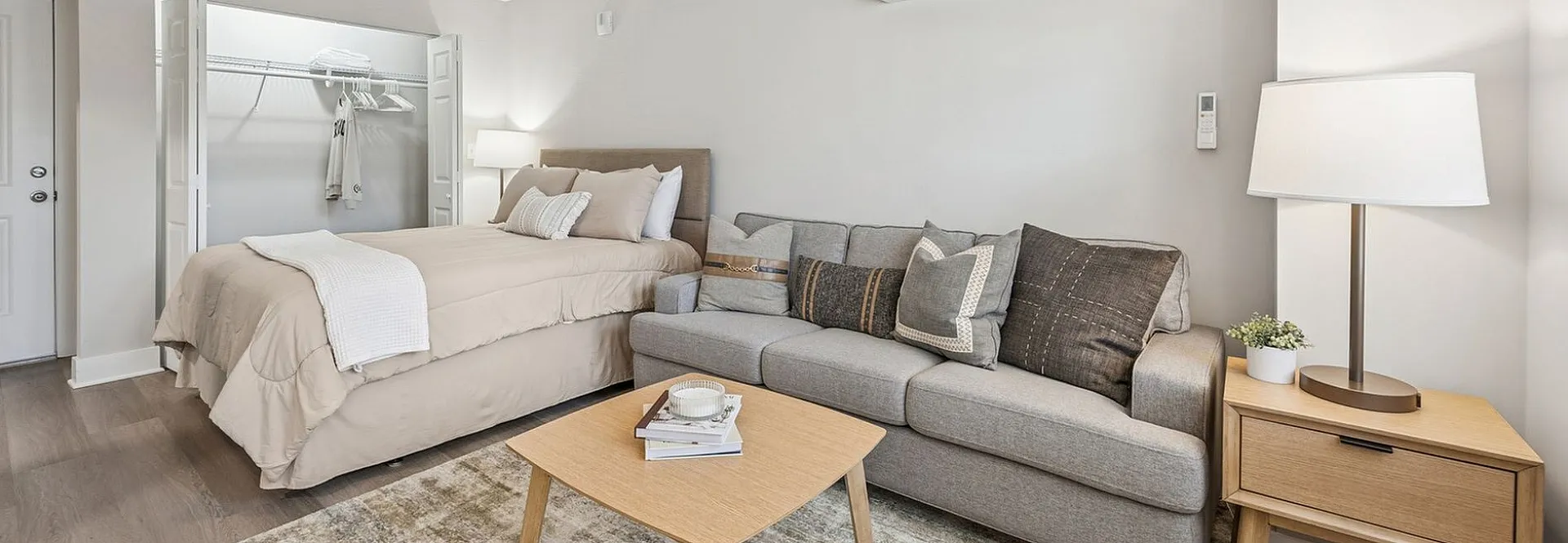 Contemporary studio apartment with elegant neutral tones, featuring a cozy bed, sleek sofa, stylish coffee table, ceiling fan, and efficient air conditioning.