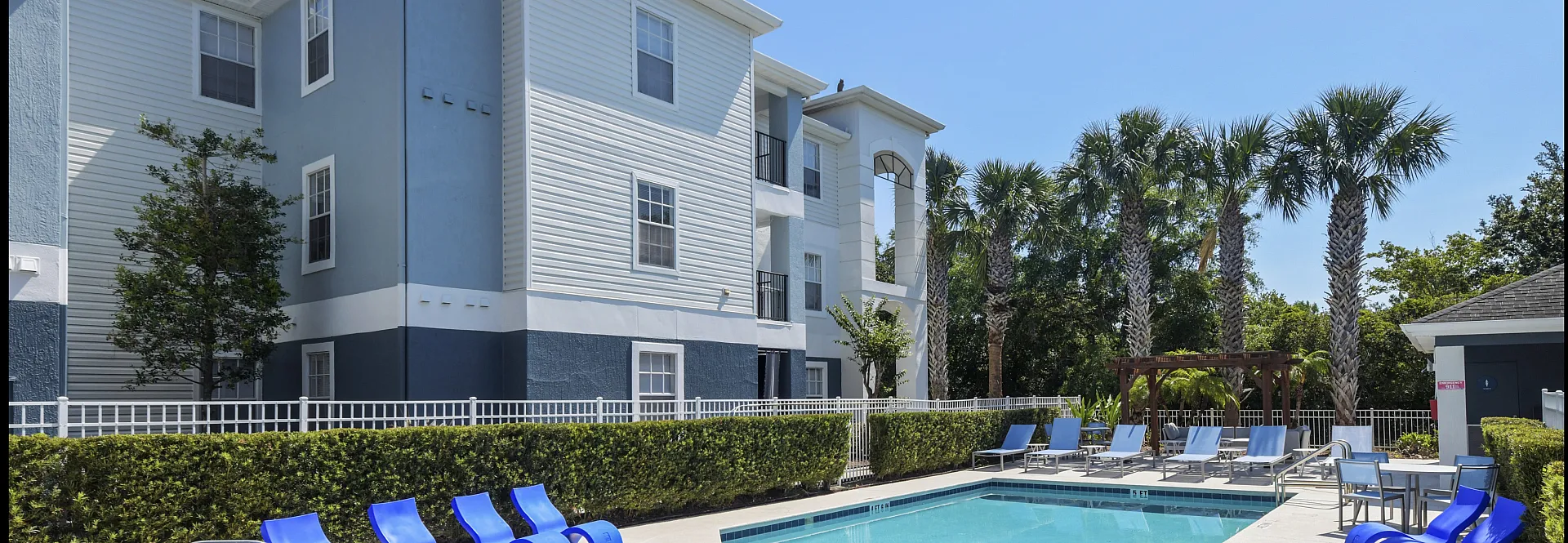 Inviting swimming pool area with stylish blue loungers, surrounded by lush palm trees and a modern apartment building backdrop.