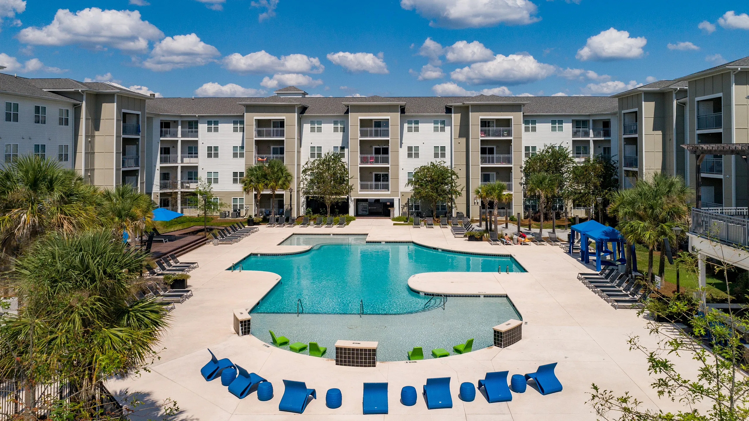 Luxurious apartment complex with a stunning resort-style pool, surrounded by lush greenery and modern architecture under a clear blue sky.