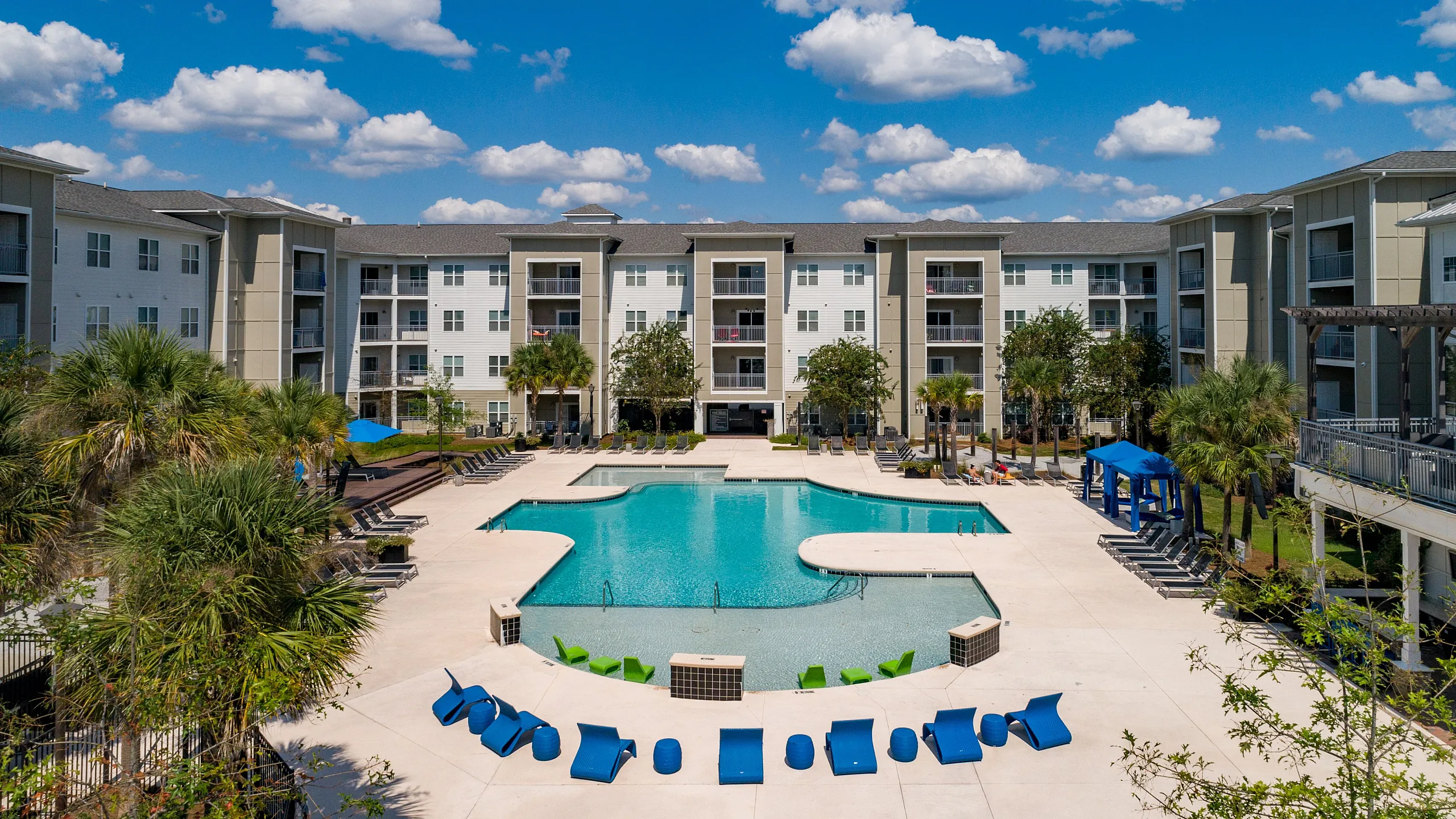 Luxurious apartment complex with a sparkling resort-style pool, surrounded by lush palm trees and modern amenities under a vibrant blue sky.