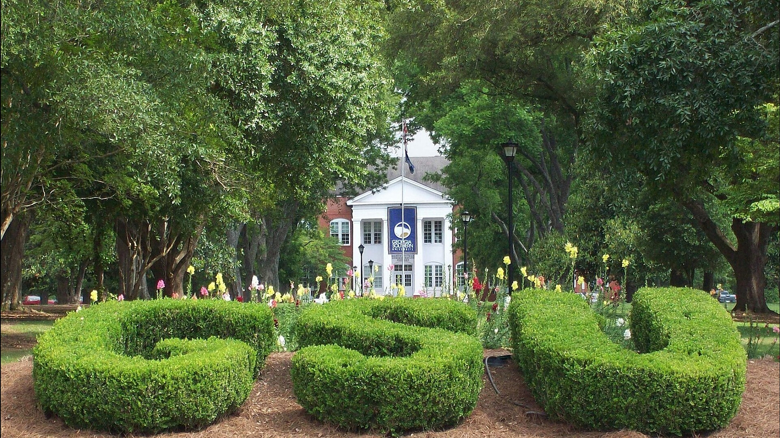 Charming campus entrance framed by lush green topiary spelling out 'GSU,' leading to a classic white-columned building with vibrant flower beds in the foreground.