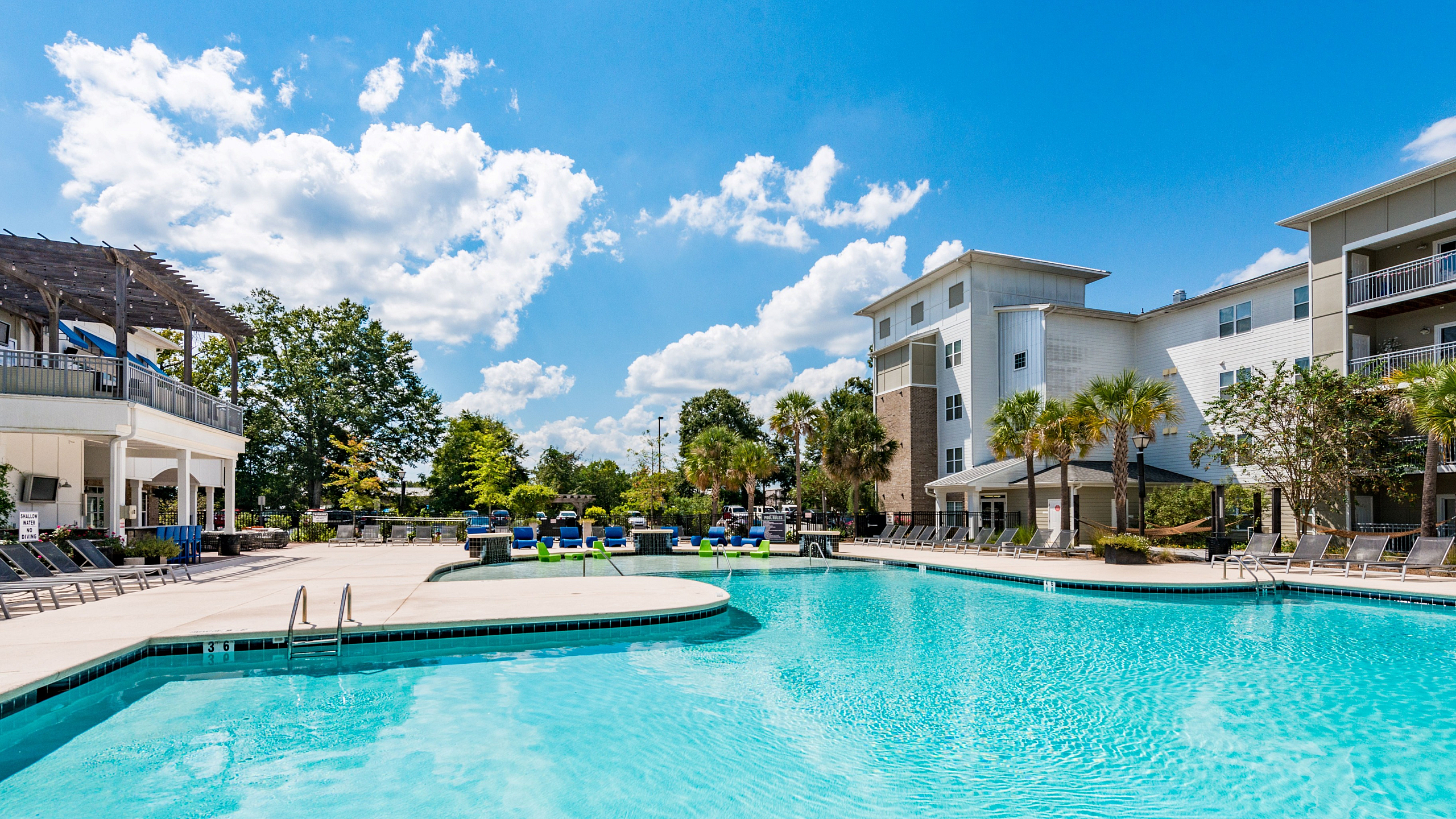 Luxurious apartment complex with a shimmering pool, surrounded by lush greenery and stylish lounging areas under a bright blue sky.