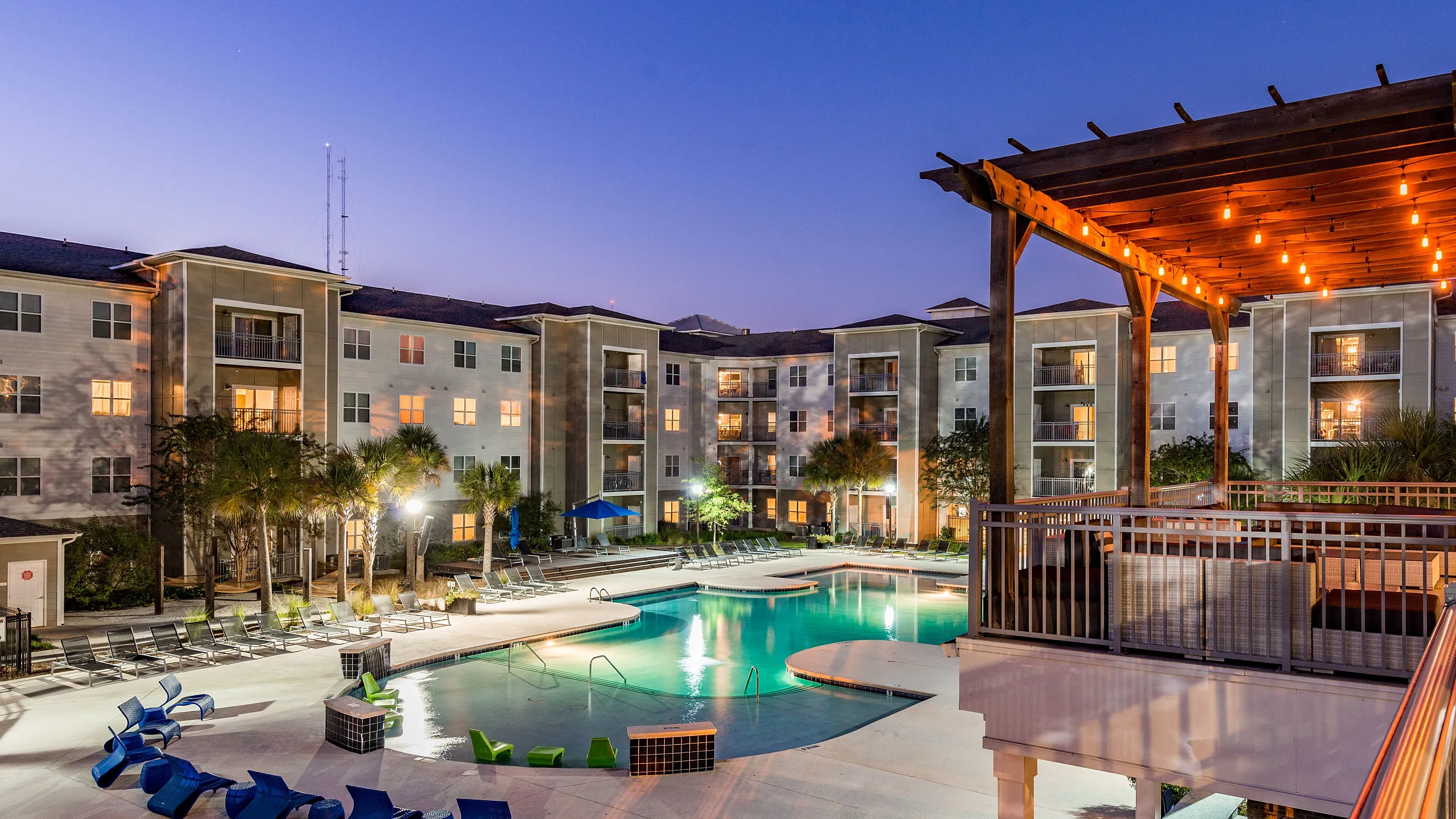 Elegant apartment complex at dusk with a beautifully illuminated courtyard and enticing swimming pool, surrounded by lush palm trees and cozy seating areas.