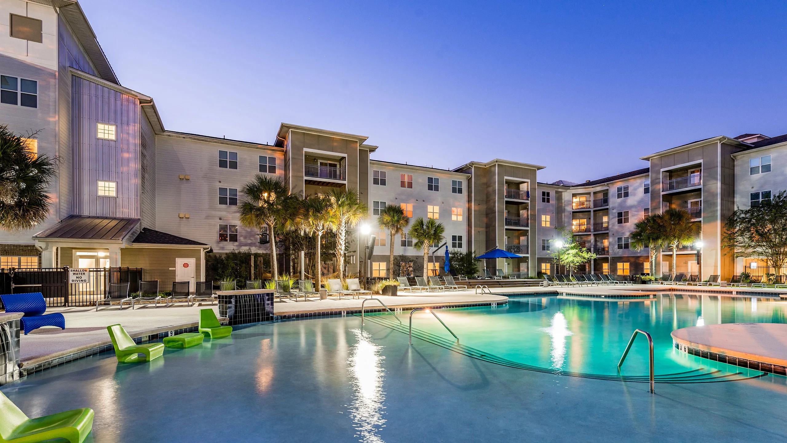 Luxurious apartment complex at dusk featuring a beautifully illuminated pool, modern architecture, and inviting lounge chairs for relaxation.