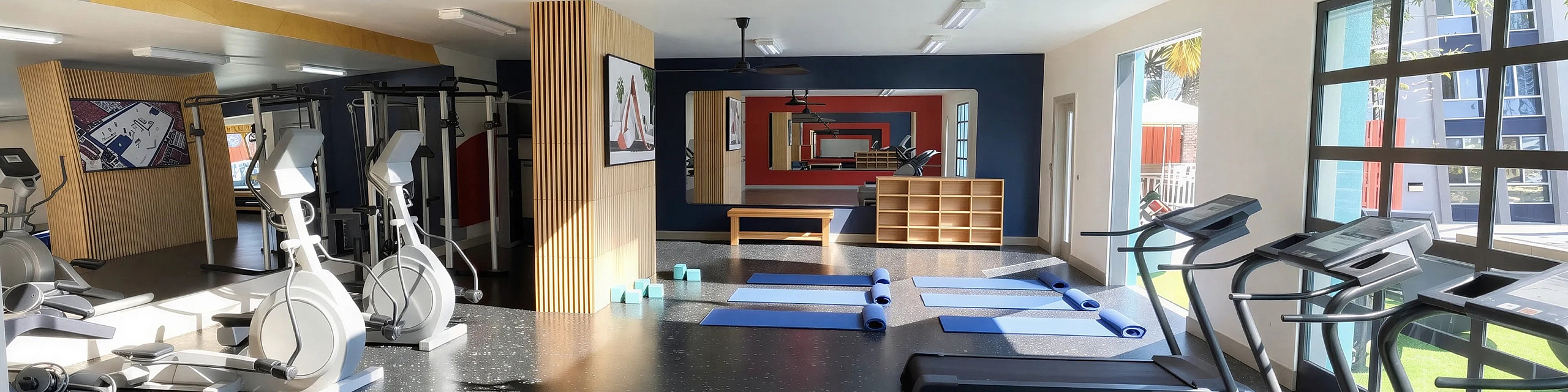Modern fitness center with state-of-the-art equipment, including treadmills, cross-trainers, and yoga mats, in a bright, sunlit space.