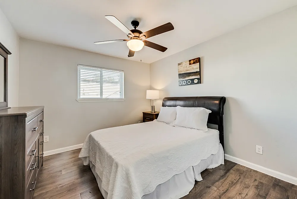 Bright bedroom with modern ceiling fan, plush bedding, and elegant hardwood floors, offering a cozy and inviting atmosphere.