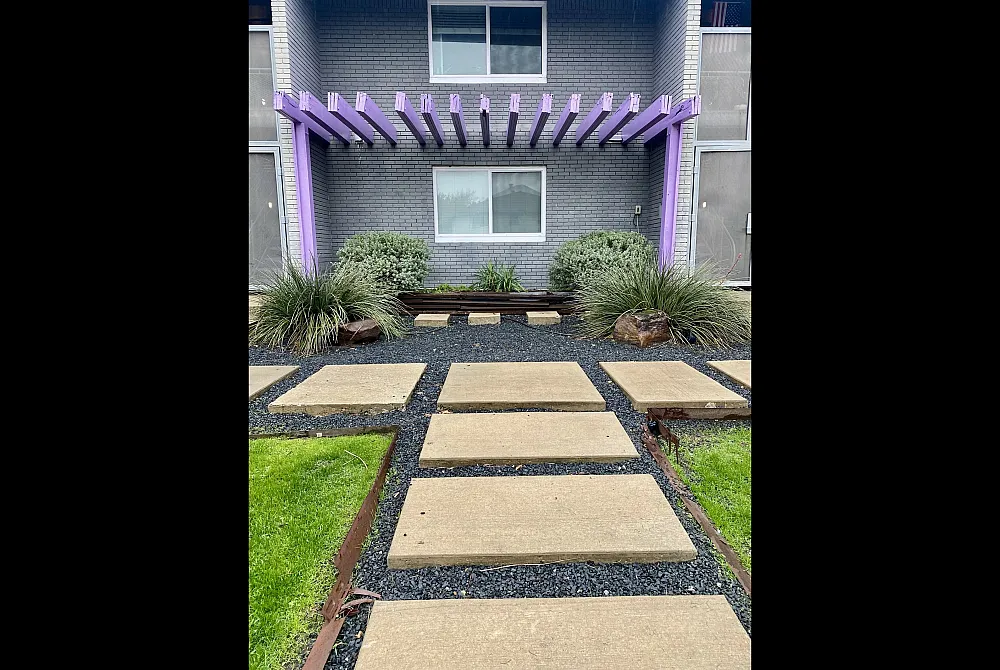 Charming apartment entrance with a modern pergola, lush greenery, and sleek stone pathway.
