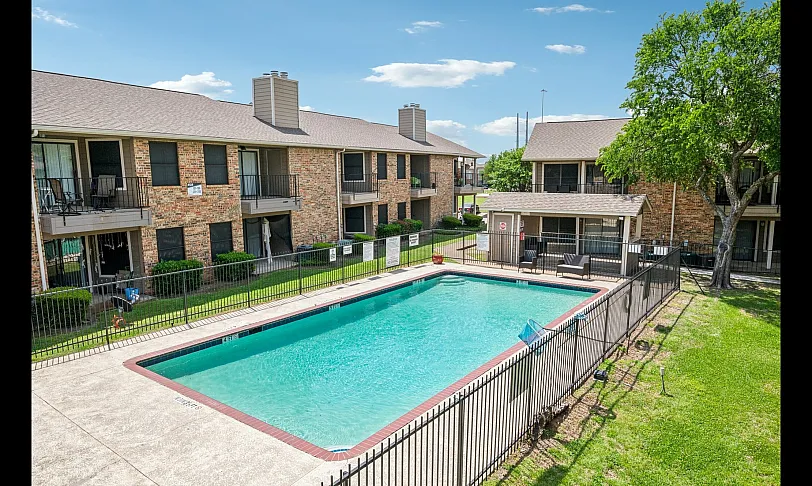 Charming apartment community with a sparkling outdoor pool, surrounded by lush greenery and brick buildings.