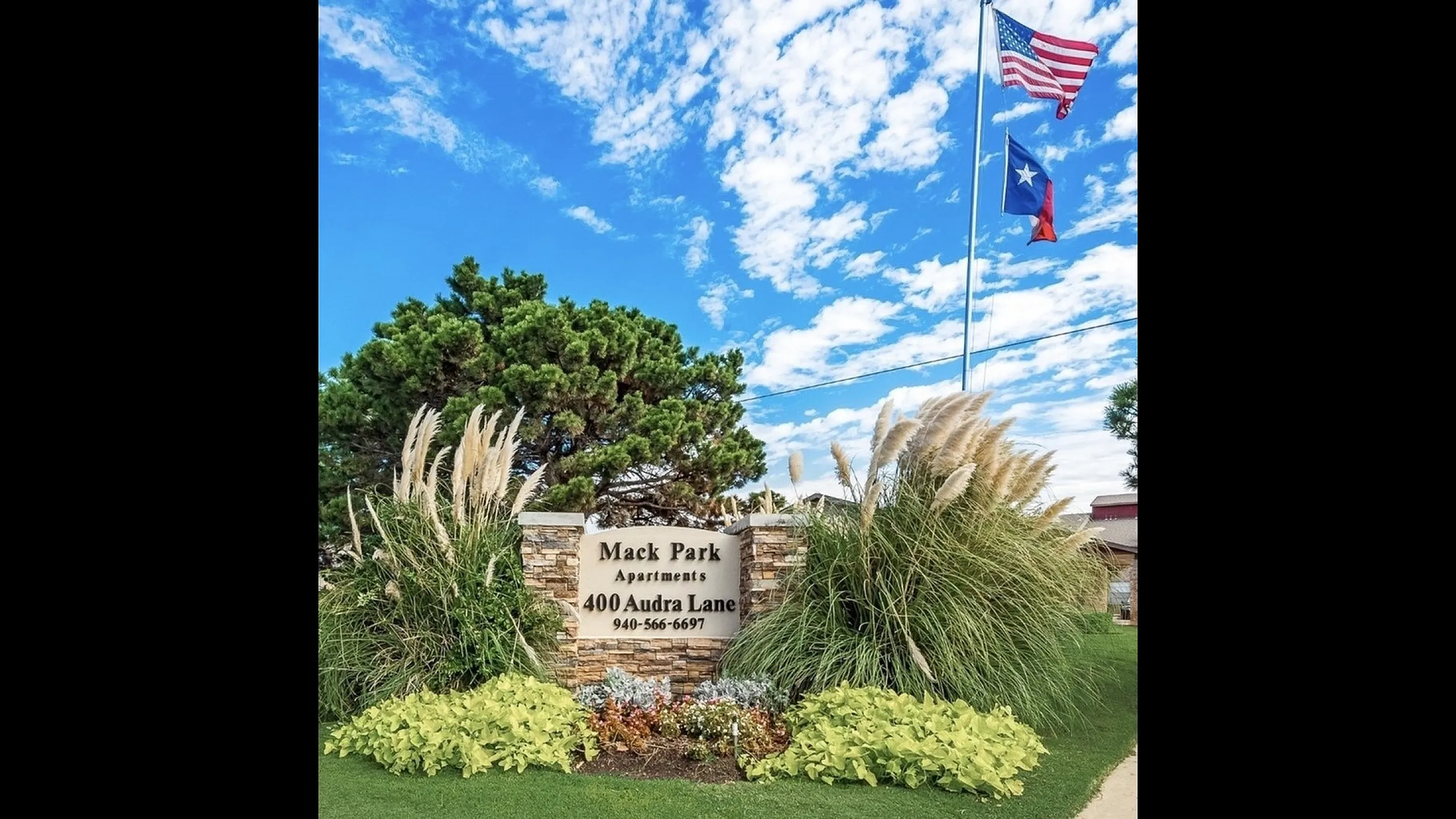 Lush entrance to Mack Park Apartments with vibrant landscaping and flags against a bright blue sky.