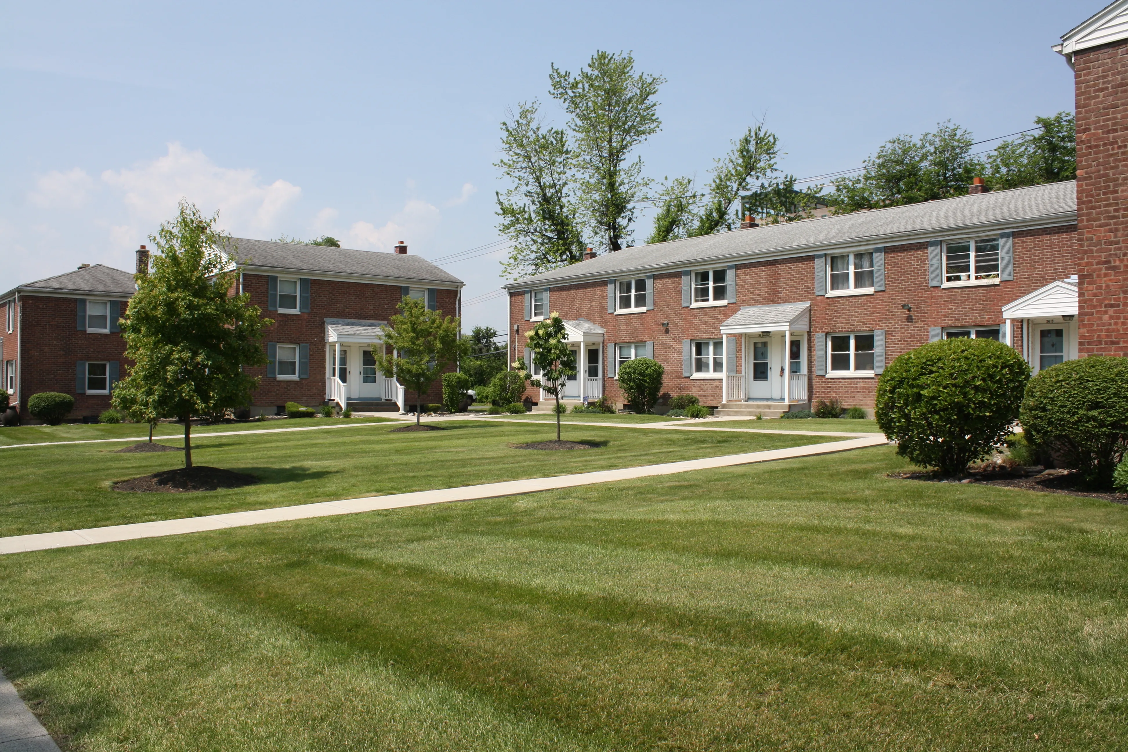 Charming brick apartment buildings on a sunny day, surrounded by lush green lawns and manicured shrubbery, nestled in a serene neighborhood setting.