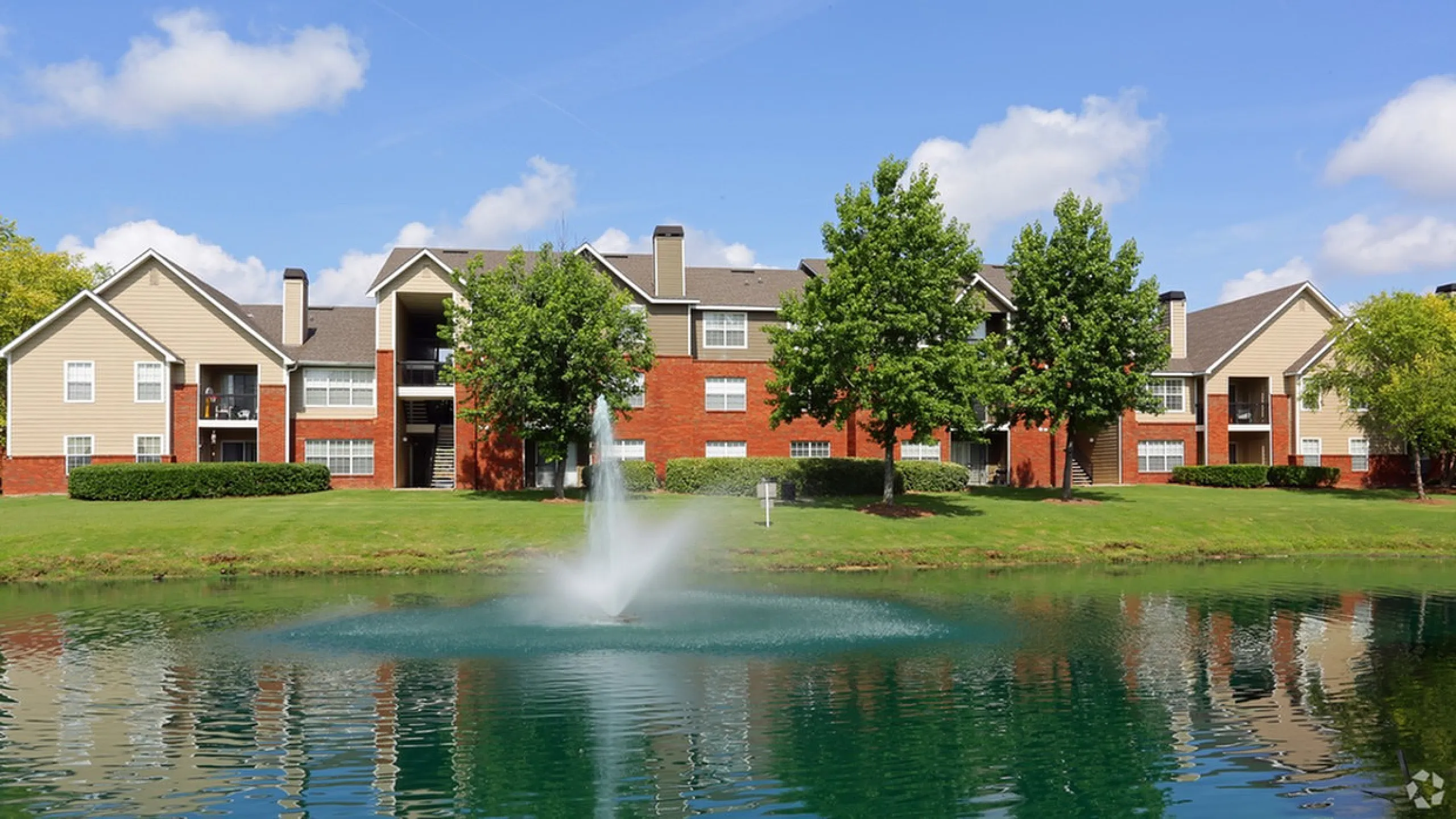 Beautiful apartment complex with a scenic pond and fountain, surrounded by lush greenery and clear blue skies.