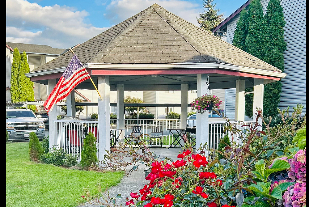 Charming gazebo with American flag surrounded by vibrant flowers, ideal for relaxation in a picturesque neighborhood setting.
