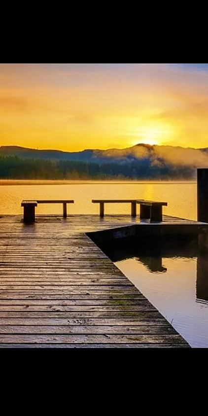 Tranquil lakeside view with a wooden pier surrounded by misty mountains and a vibrant sunset sky.