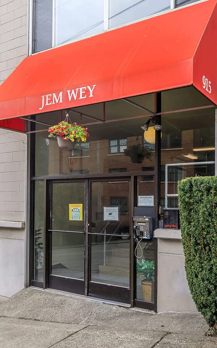Inviting apartment entrance with a vibrant red awning and elegant glass doors, framed by neatly trimmed shrubbery and colorful hanging flowers.