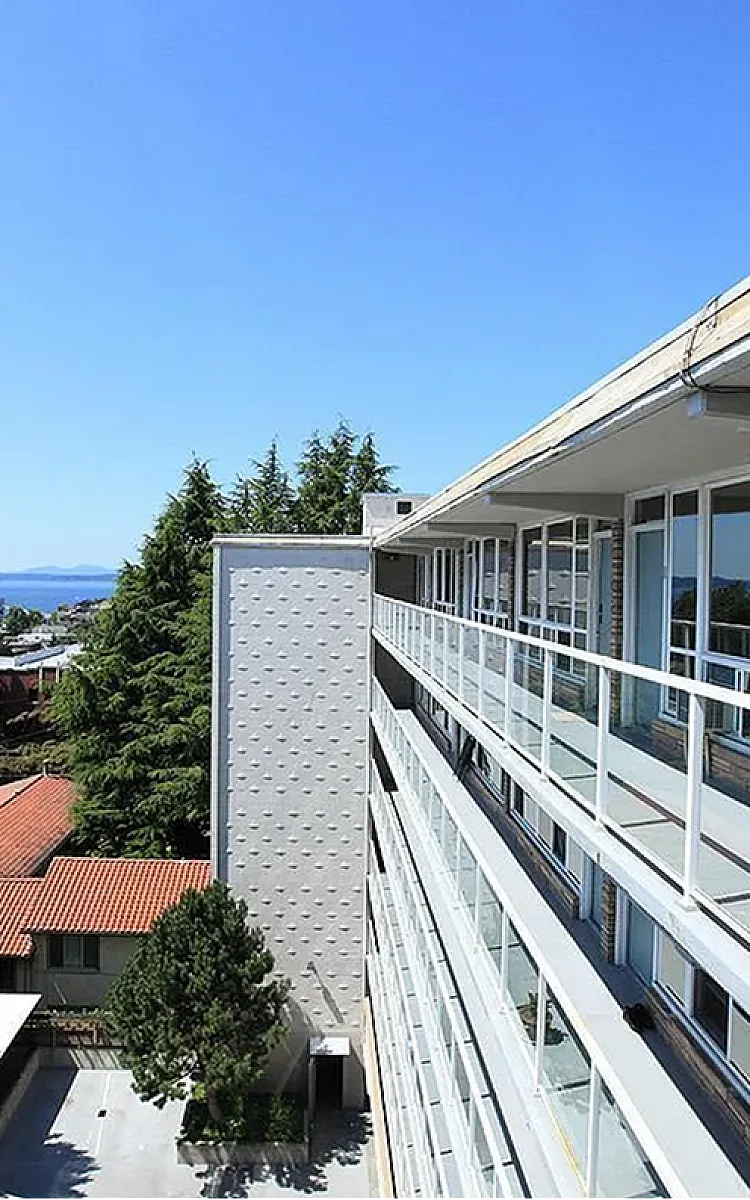 Sleek apartment building with panoramic ocean views from a spacious balcony, surrounded by lush greenery and clear blue skies.