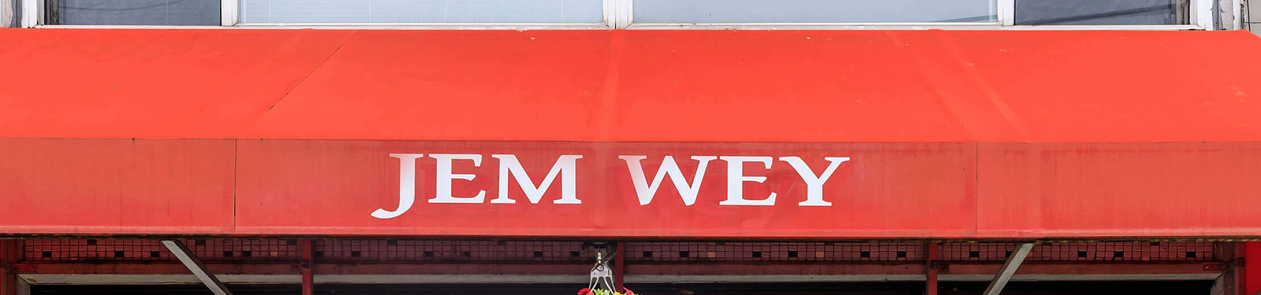 Charming storefront with a bright red canopy displaying 'Jem Wey' and a vibrant hanging flower basket, offering a welcoming entrance.