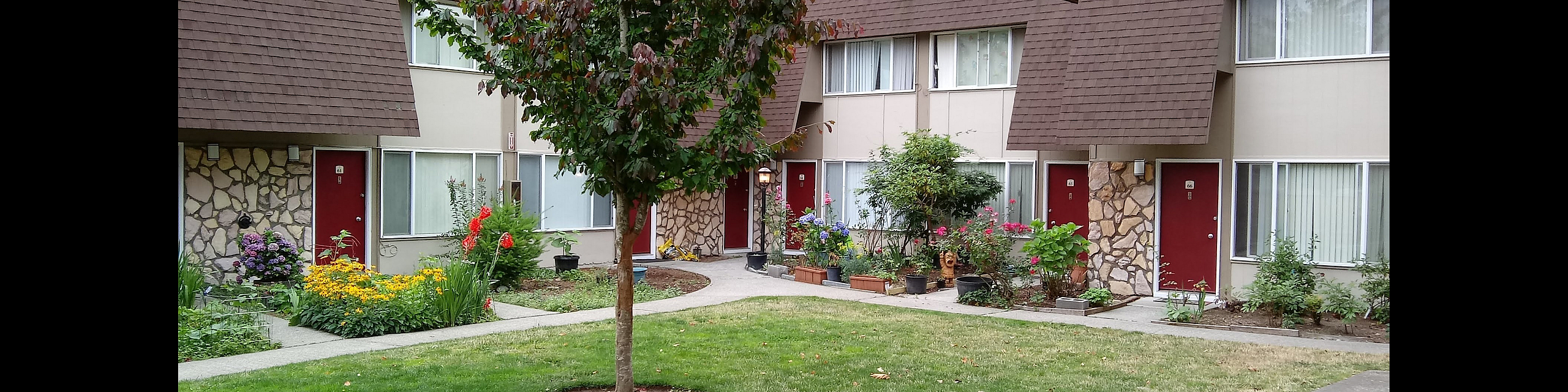 Charming apartment complex with cozy brown roofs and vibrant gardens, surrounded by lush greenery and a serene pathway.