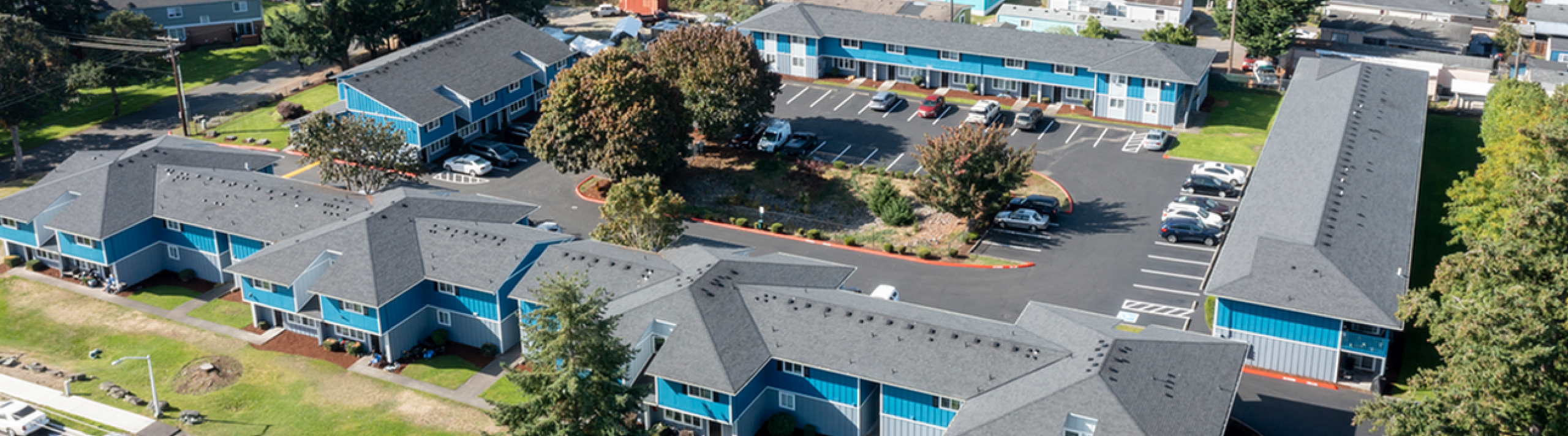 Charming aerial view of a modern apartment complex nestled in a lush green neighborhood, featuring vibrant blue buildings with ample parking and scenic surroundings.