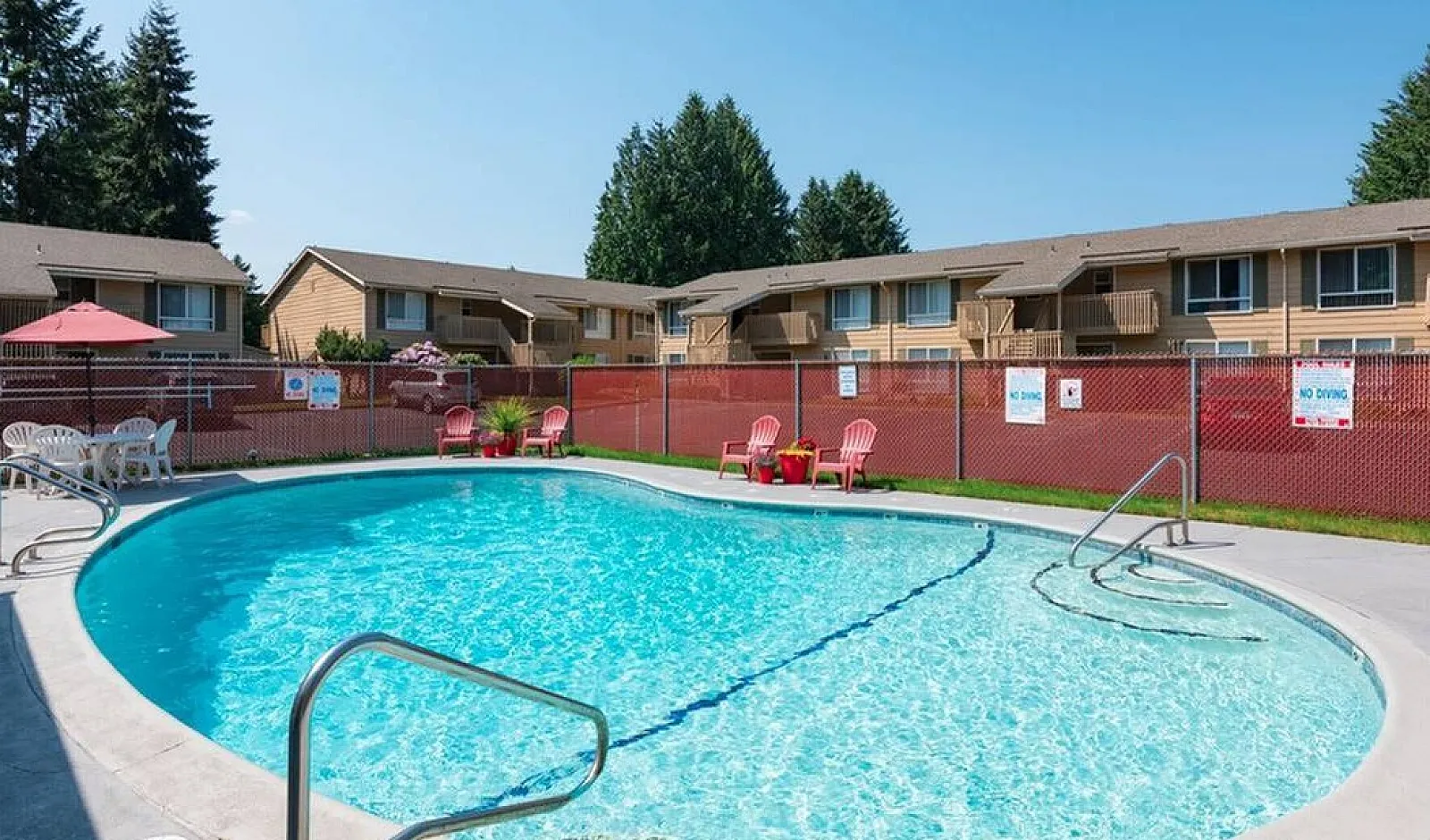 Charming apartment community with a sparkling pool, relaxing lounge chairs, and vibrant landscaping under a clear blue sky.