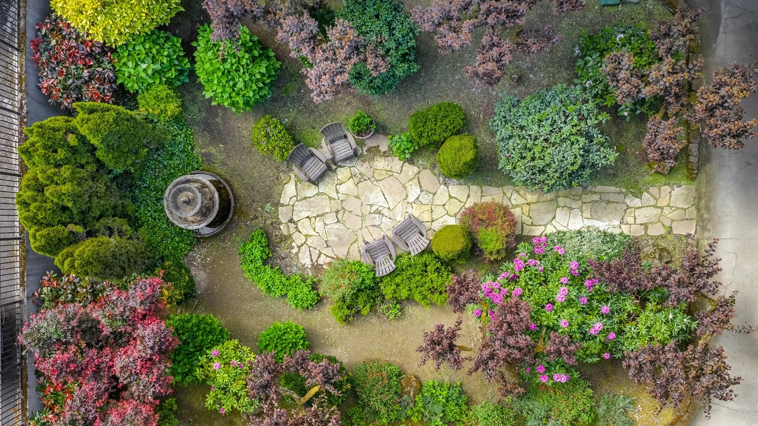 Charming aerial view of a lush garden oasis with vibrant flowering plants, cozy seating area, and a centerpiece stone fountain.