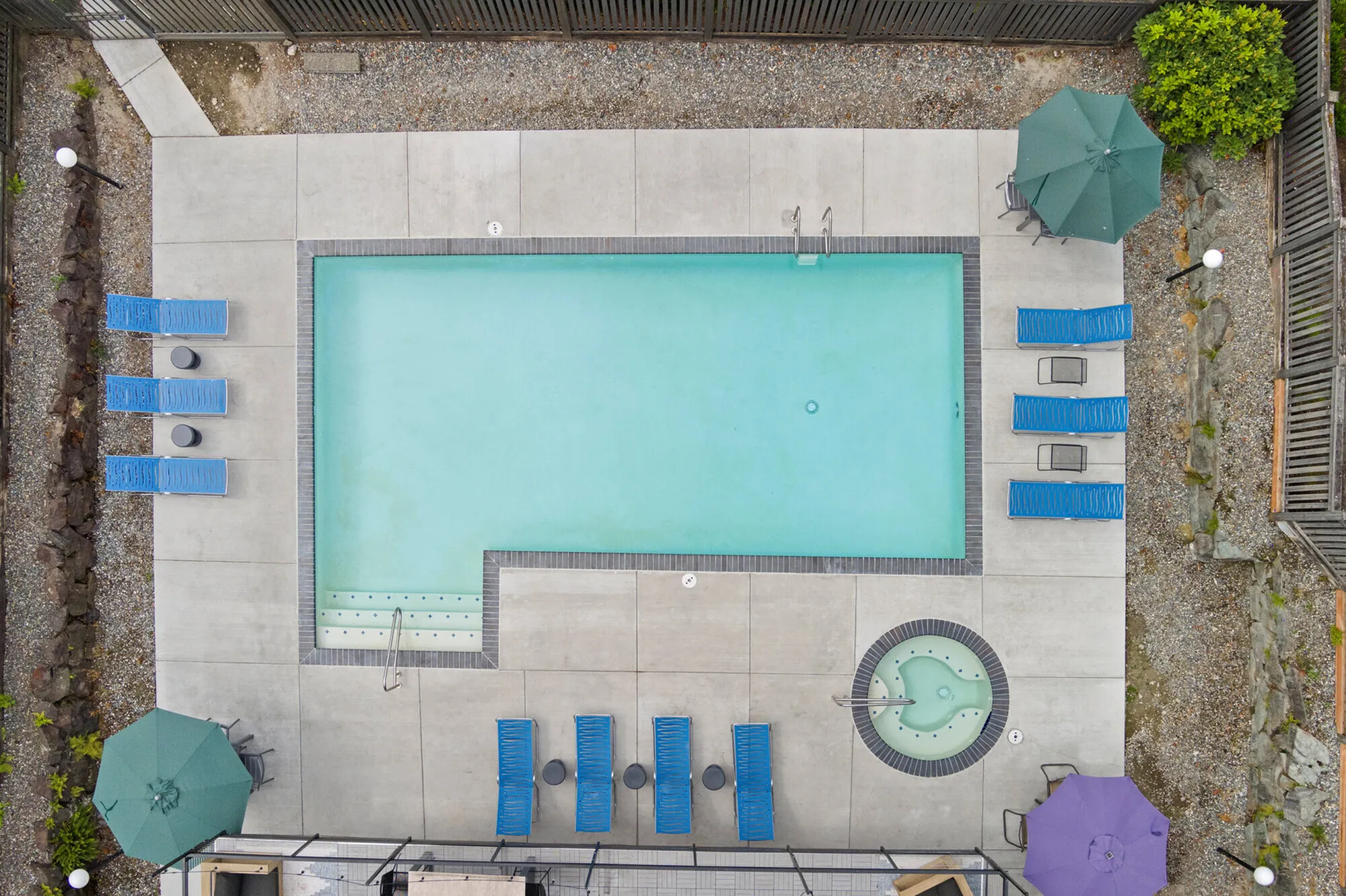 Inviting aerial view of a modern apartment pool area with vibrant blue lounge chairs, umbrellas, and a cozy hot tub, perfect for relaxing in style.