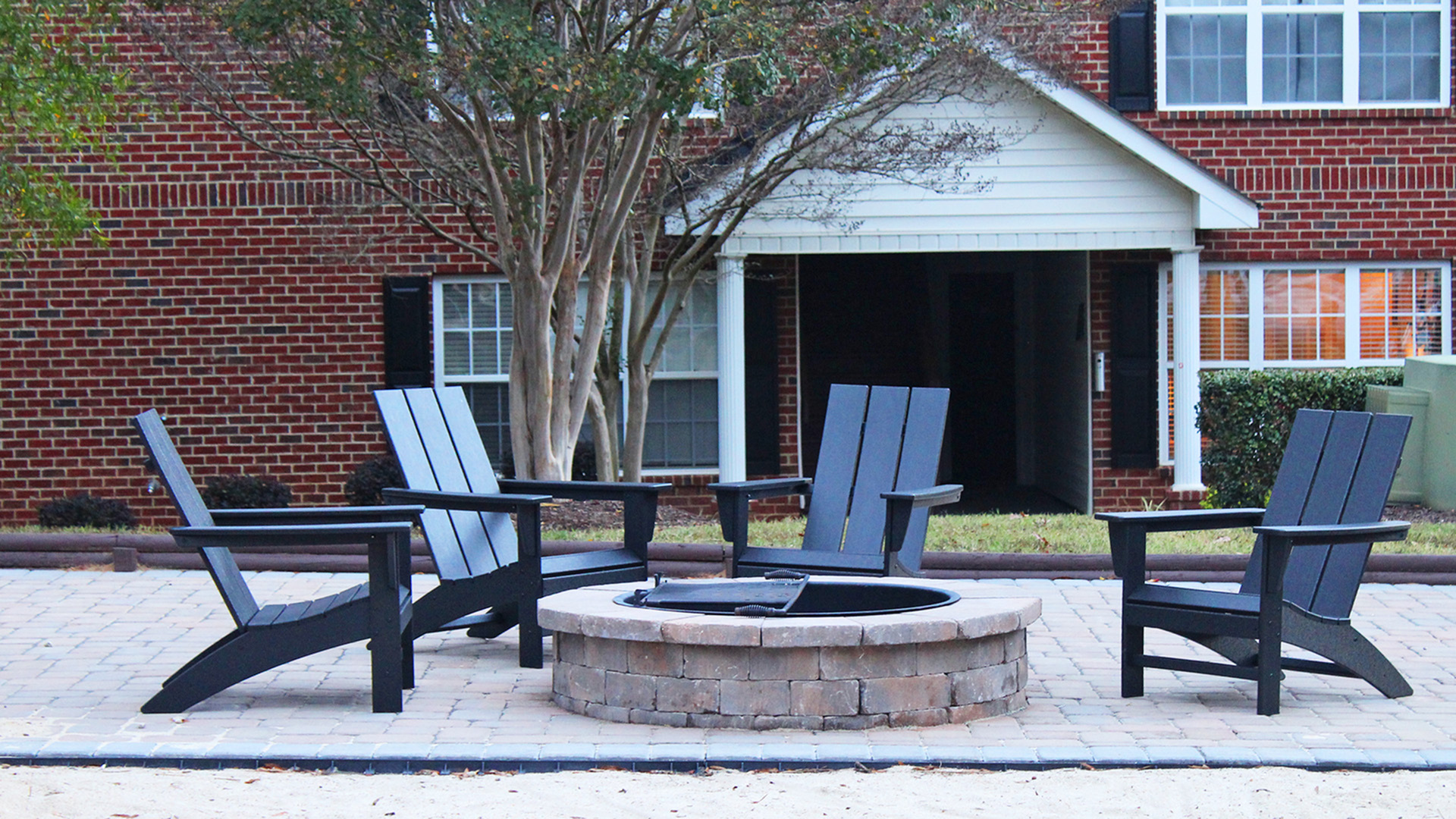 Inviting outdoor seating arrangement featuring four Adirondack chairs around a stone fire pit, set against a charming brick building.