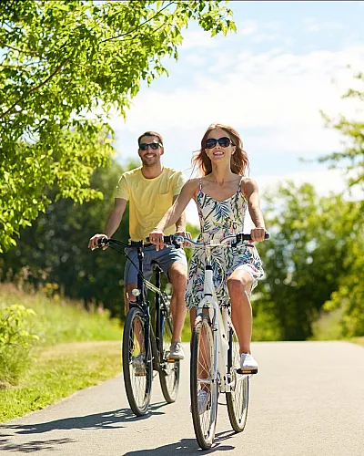 Joyful couple cycling on a scenic path lined with lush green trees under a bright blue sky.