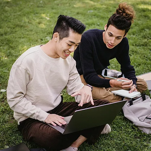 Two smiling individuals enjoy a productive outdoor work session on a laptop in a green park setting, promoting a relaxed and collaborative atmosphere.