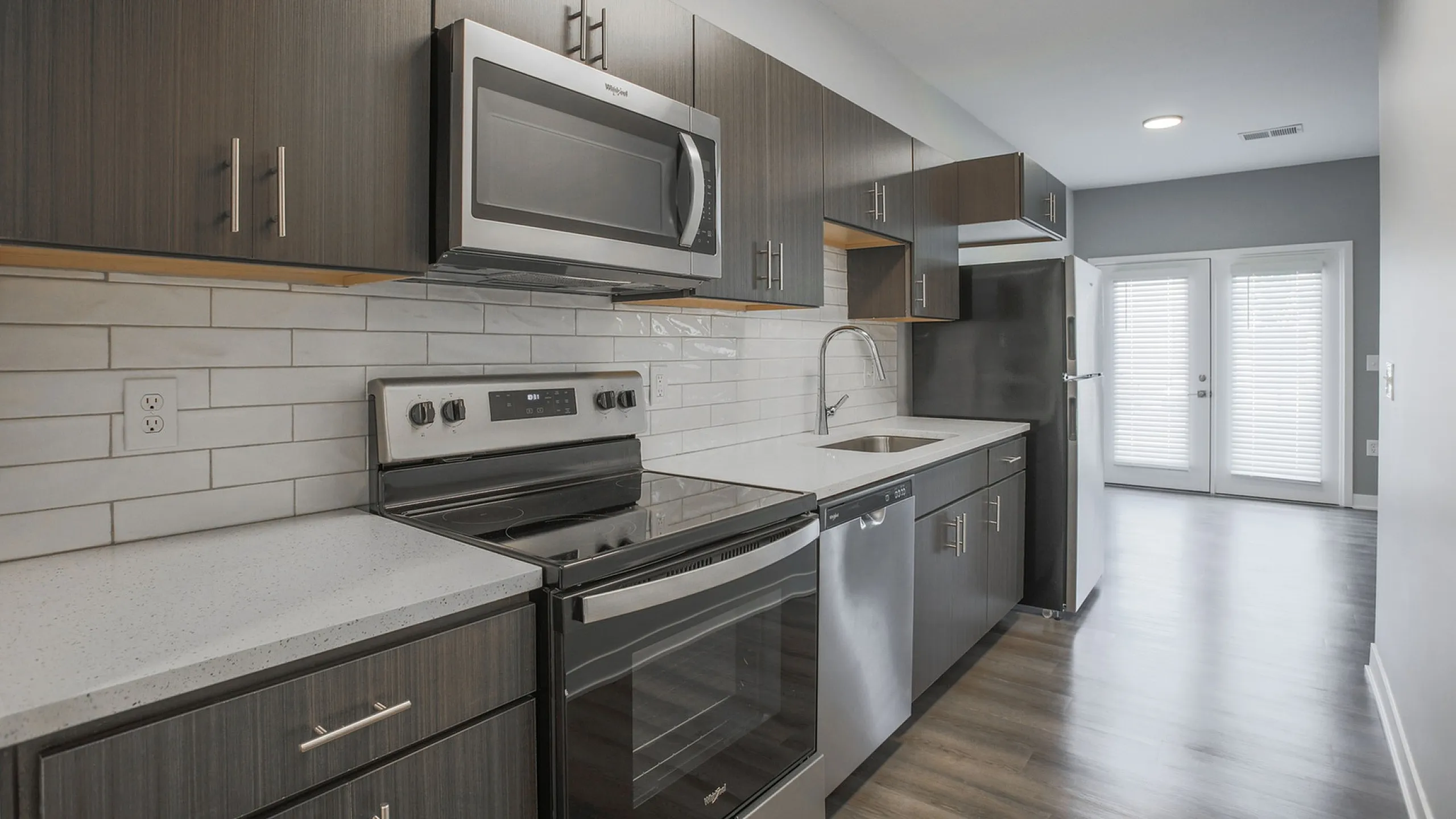 Modern kitchen with dark wood cabinets, stainless steel appliances, and sleek countertops, leading to a bright living area with natural light.