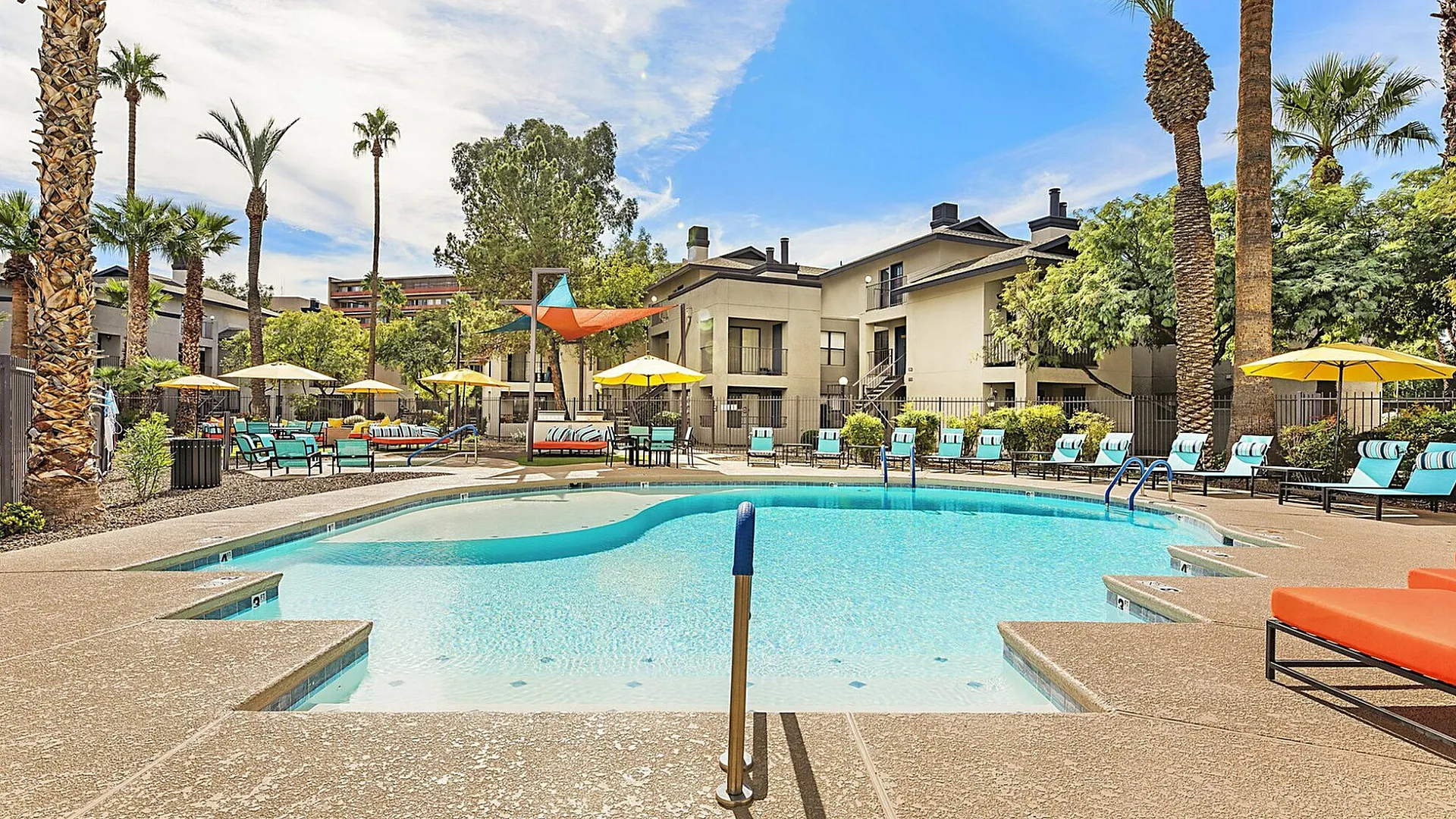 Inviting apartment complex pool area with vibrant sun loungers, lush palm trees, and sunny ambiance ideal for relaxation.