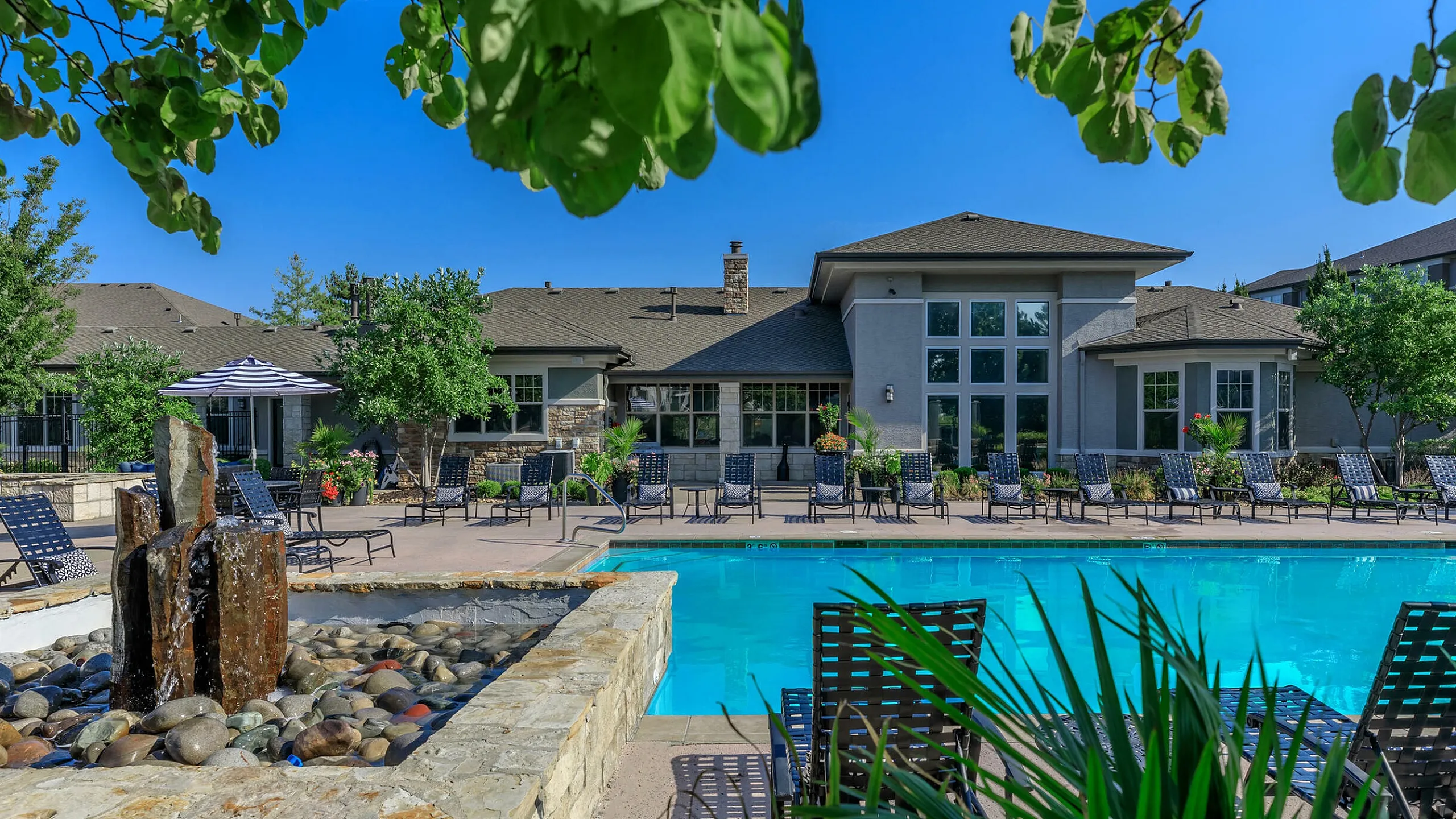 Corbin Crossing luxurious apartment complex showcases a serene pool area with lounge chairs and elegant stone fountain, surrounded by lush greenery.