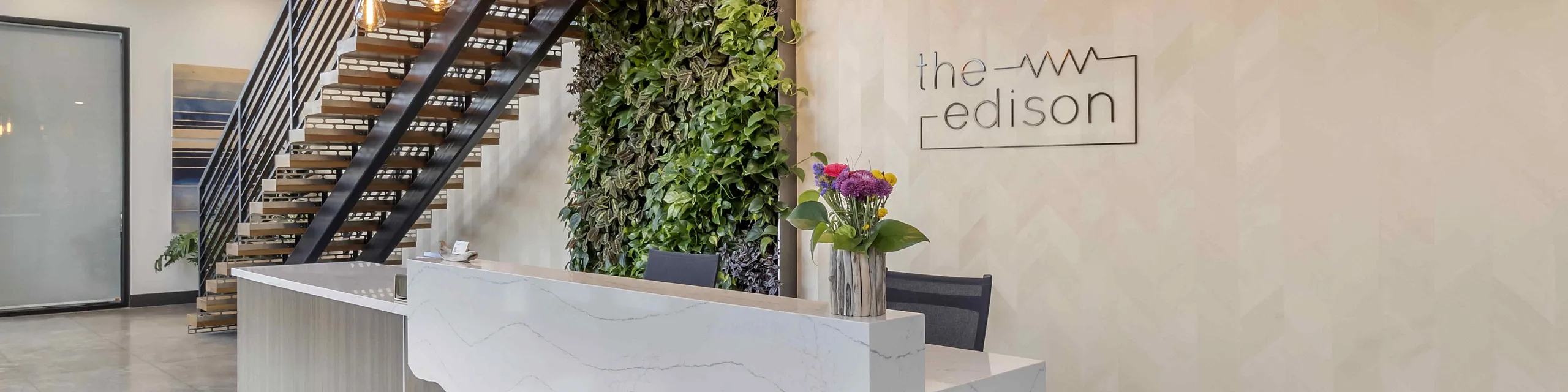 Modern apartment lobby featuring a sleek reception desk, elegant staircase, lush green vertical garden, and stylish lighting, offering a warm welcome to the Edison.