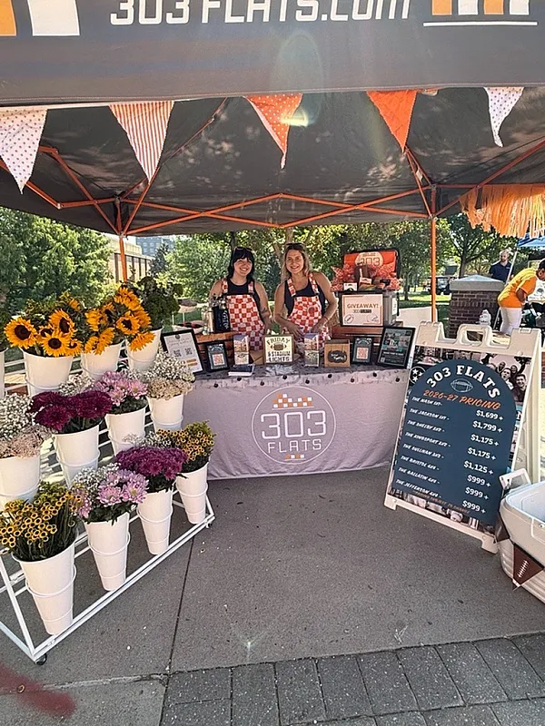 Inviting outdoor booth for 303 Flats featuring vibrant flowers, cheerful hosts in checked aprons, and enticing promotional materials under a colorful canopy.