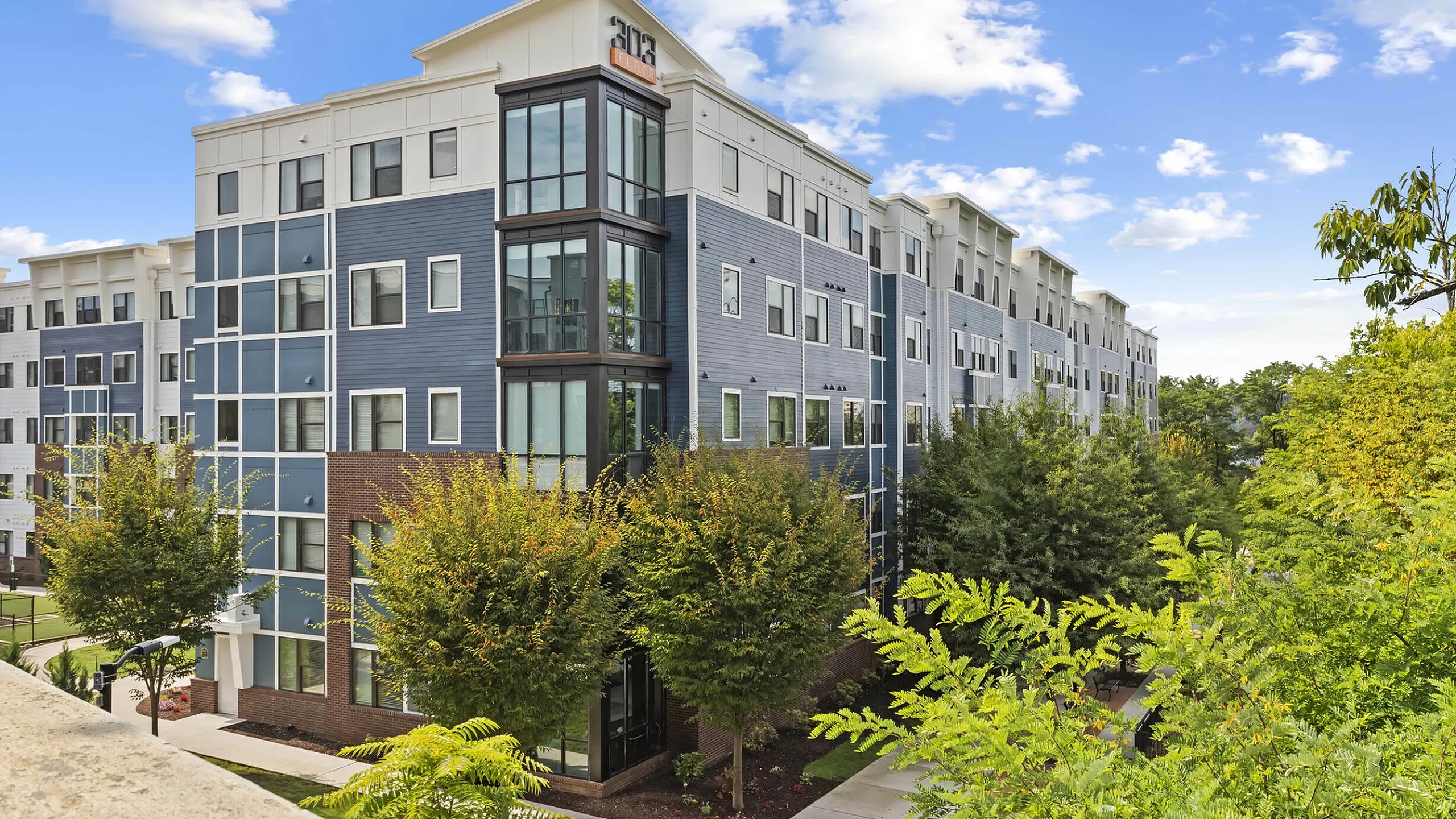 303 Flats apartment building with stylish architectural design, surrounded by lush greenery and vibrant trees, under a clear blue sky.